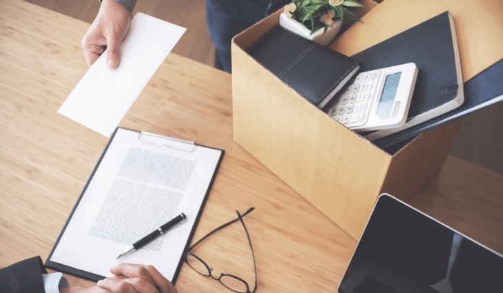 A person hands over a letter; a box of personal belongings sits nearby on a desk.
