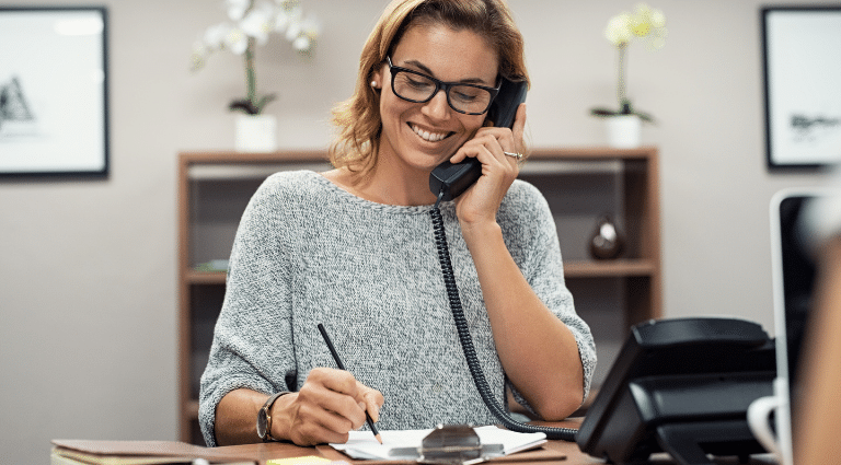 Smiling woman in glasses takes notes while on the phone in an office setting.