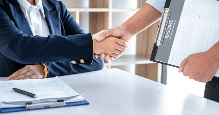 A professional handshake between two people, a resume on a clipboard present.