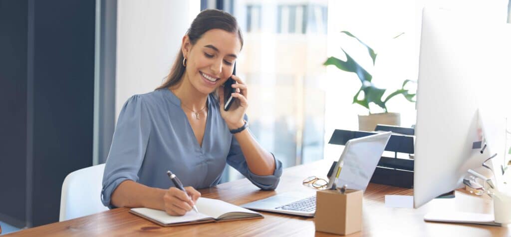 A smiling woman in a blue top sits at a desk taking notes while on a phone call with a laptop and desktop computer nearby