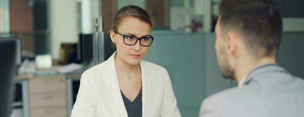 Professional woman in a white blazer and glasses listens to a male colleague in an office setting, mid-conversation.