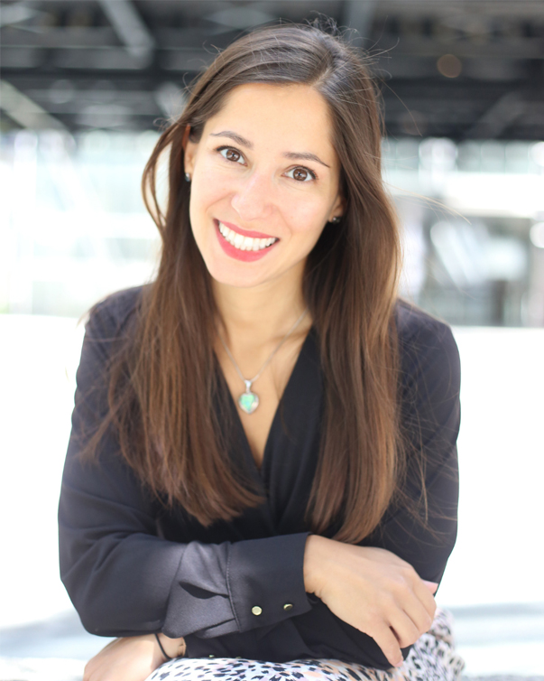 Smiling woman with long brown hair in a black blouse leaning forward outdoors in bright natural light wearing a turquoise pendant necklace