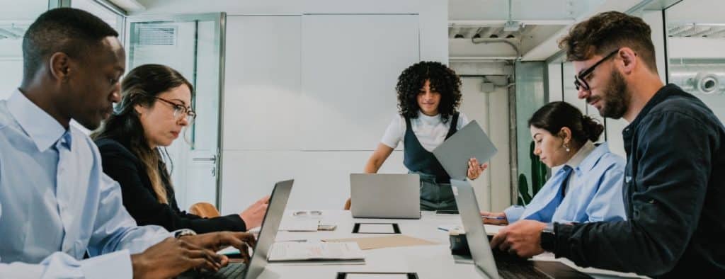 Team of five professionals around a conference table with laptops, while one woman stands presenting with a folder in a bright office.