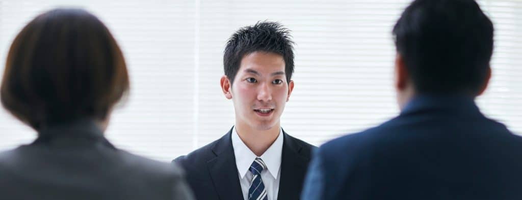 Young man in a suit explains something to two colleagues in a bright office meeting room.